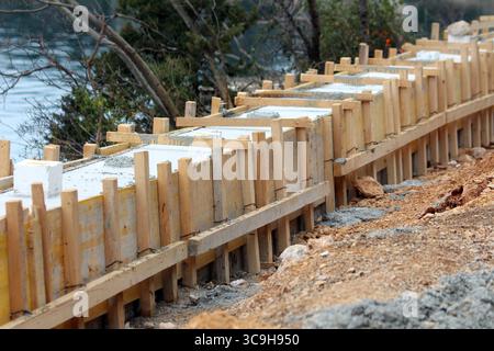 New concrete is setting inside sturdy wooden formwork, creating a straight foundation line for a riverside path, surrounded by soil and sparse Stock Photo