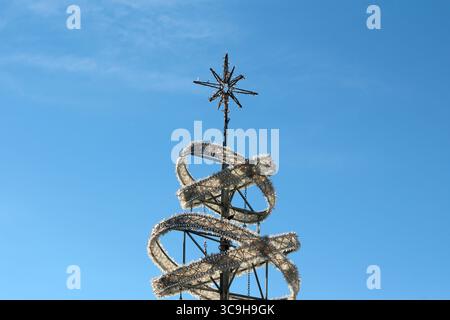 A metallic sculpture features a spiraling white garland and a star topper, set sharply against a vivid blue sky, evoking a contemporary holiday Stock Photo
