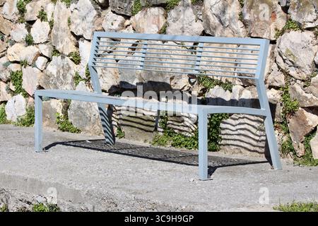 A sleek, light gray metal bench stands on concrete, casting bold striped shadows against a rustic stone wall with scattered green ivy and moss Stock Photo