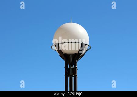 A contemporary white globe streetlamp is mounted on a black metal frame, standing tall and isolated against a clear, vibrant blue sky Stock Photo