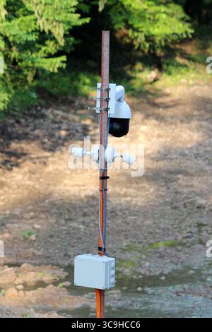 Multiple security cameras and electrical box are installed on a rusty pole, monitoring a forest clearing with dense trees in the background Stock Photo