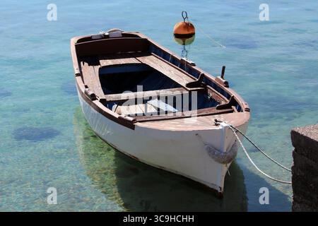 A rustic wooden rowboat with peeling paint floats gently in transparent turquoise water, tethered near a concrete dock with a visible orange buoy Stock Photo