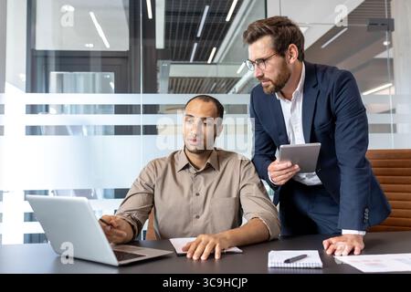 Two business professionals collaborating in an office setting, discussing data on a laptop and tablet. Stock Photo