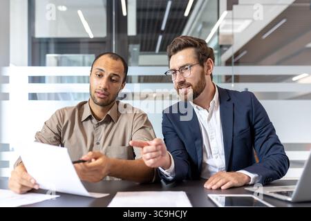 Two diverse businessmen collaborate, reviewing documents at a modern office desk, discussing and analyzing data for a project. Stock Photo
