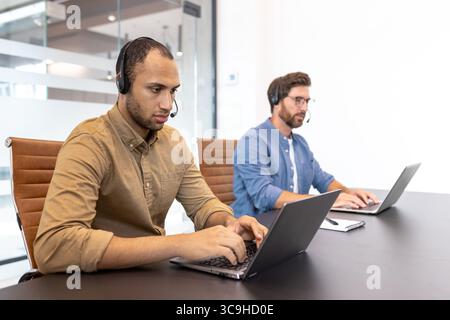 Two diverse call center agents work on laptops, wearing headsets in a modern office environment, providing customer support. Stock Photo