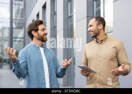 Two diverse businessmen are having a lively conversation outdoors in front of a modern building, possibly discussing a project or deal. Stock Photo