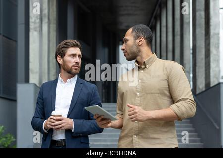 Two business professionals discuss work while looking at a tablet outside a modern building. One holds a coffee cup. Stock Photo