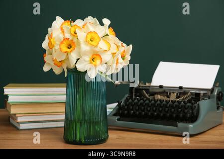 Vase with yellow flowers, retro typewriter and books on wooden table against green background Stock Photo