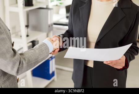 Two businesswomen in formal suits holding contract, making agreement and shaking hands in office. Stock Photo