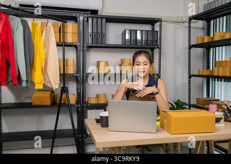 Young female entrepreneur working at home, managing her online clothing store business, checking orders from customers via laptop and preparing packag Stock Photo