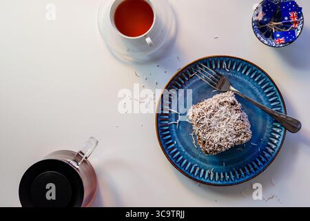 A Lamington cake with two Australian flags, one on the cake and one on a teacup saucer. A fork, blue plate, and glass teapot are on the white table. Stock Photo
