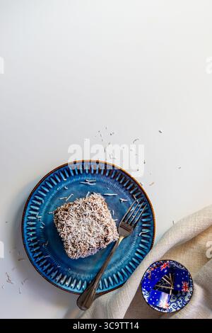 A Lamington cake with two Australian flags, one on the cake and one on a teacup saucer. A fork, blue plate, and glass teapot are on the white table. Stock Photo