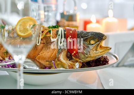A beautifully presented roasted fish dish garnished with lemon and colorful vegetables, set elegantly on a dining table surrounded by soft candlelight Stock Photo