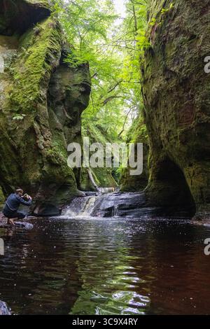 Small cascading waterfalls nestled between moss-covered cliffs in a lush, green forest gorge. The tranquil water flows through a dramatic Stock Photo