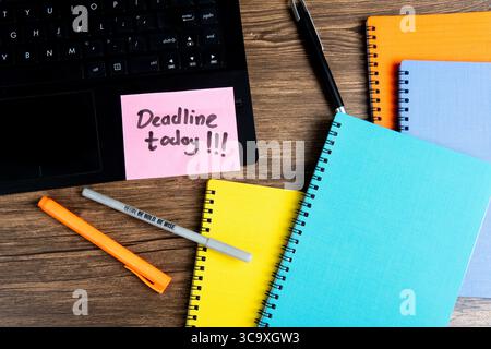 Flat lay of colorful notebooks, pens, and laptop on wooden desk with a sticky note reminder saying "Deadline today!!!". Stock Photo