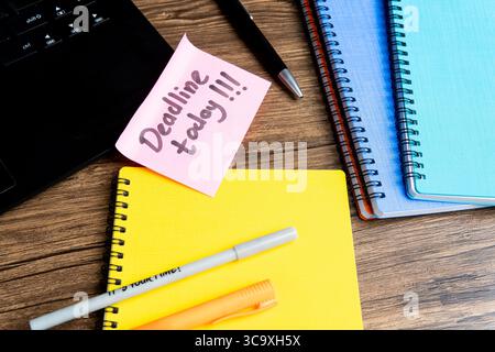 Flat lay of colorful notebooks, pens, and laptop on wooden desk with a sticky note reminder saying "Deadline today!!!". Stock Photo