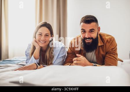 Young happy couple is lying on bed in their bedroom, looking at camera and smiling. Stock Photo