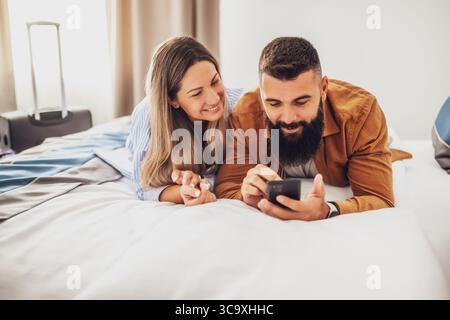 Young happy couple is on vacation. Tourists arrived in hotel room with their suitcases. They are lying on bed and using smartphone. Stock Photo