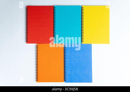 Top view of five spiral notebooks in red, orange, blue, yellow, and teal. Arranged on white textured surface. Stock Photo
