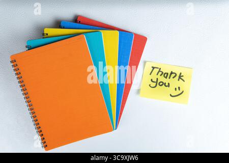 Close-up of colorful folders and a yellow sticky note with handwritten text "Thank You" and smiley face on a white wall. Stock Photo