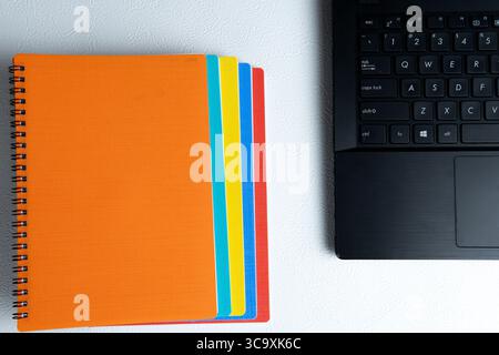 Top view of stacked colorful spiral notebooks and part of a black laptop on a white textured desk. Stock Photo