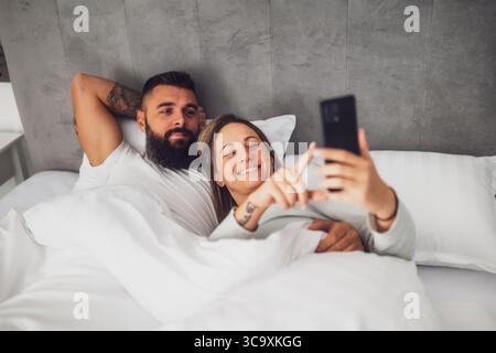Young happy loving couple is lying in bed in the morning. They are relaxing and using smartphone. Stock Photo