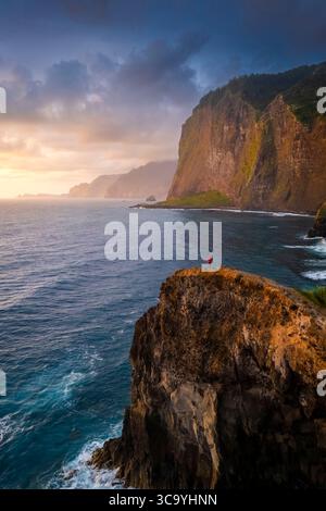 Aerial view of a tourist admiring sunrise from the cliffs of Miradouro do Guindaste. Madeira, Portugal, Europe. Stock Photo