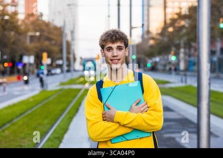 Smiling college student holding a binder and wearing backpack in urban setting Stock Photo