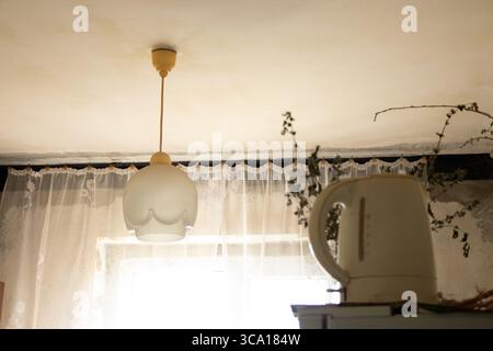 Chandelier on the ceiling and an electric kettle on the refrigerator in an old Soviet kitchen with curtains and dried branches, everyday home interior Stock Photo
