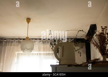 Chandelier on the ceiling and an electric kettle on the refrigerator in an old Soviet kitchen with curtains and dried branches, everyday home interior Stock Photo