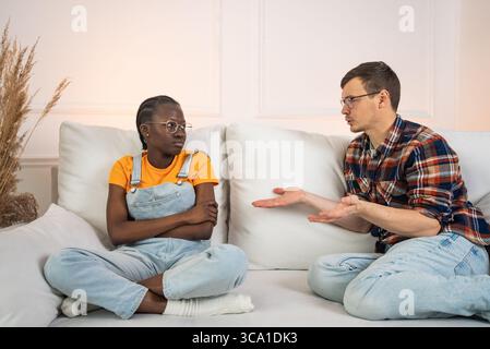 Young multiethnic couple sitting on a sofa in their living room, discussing relationship difficulties while expressing frustration and tension during a serious conversation Stock Photo