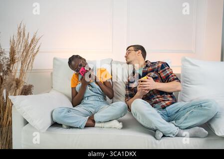 Young interracial couple sitting together on a cozy white sofa in their bright living room, with the woman recording a voice message on her smartphone while the man enjoys a cup of tea Stock Photo