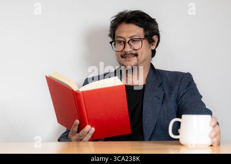 Relaxed young man wearing glasses enjoying reading a book in a cozy home environment. Stock Photo