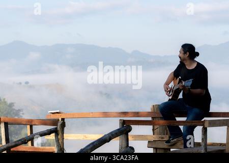Young man enjoying a peaceful morning, playing guitar on a wooden balcony with warm sunlight and scenic mountain views. Stock Photo