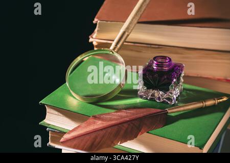 Literature concept. Inkstand with feather near magnifying glass on old books against black background. Stock Photo