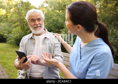 Nurse and elderly man using smartphone on bench outdoors Stock Photo