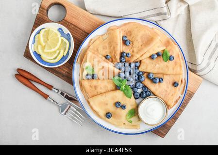 Homemade thin pancakes (crepes) with fresh blueberries and sour cream on white plate on light background, top view Stock Photo