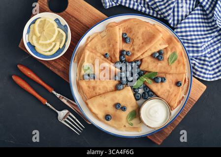 Homemade thin pancakes (crepes) with fresh blueberries and sour cream on white plate on black background, top view Stock Photo