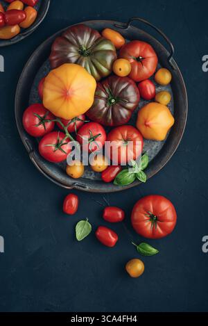 Assortment of ripe tomatoes varieties. Vintage metal tray of colorful tomatoes and cherry tomatoes on black background, top view. Stock Photo