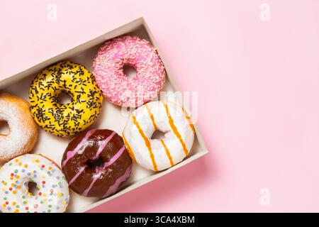 Box of six assorted donuts with colorful icing, stripes, and toppings on pastel pink background with copy space and space for text Stock Photo