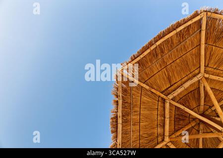 Tropical background with thatched beach umbrella seen from below in the corner and blue sky, copy space, holidays. Stock Photo