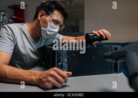 Focused computer technician in protective mask and safety eyewear cleaning desktop tower with handheld vacuum during precision hardware maintenance. Stock Photo