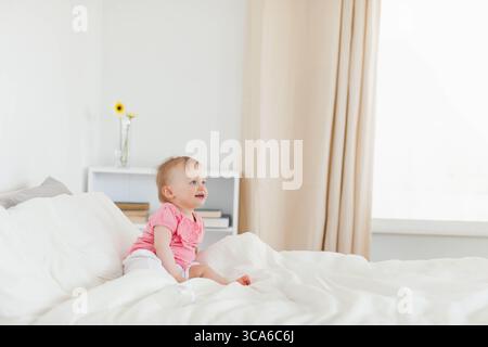 Baby girl sitting upright on bed in bedroom beside books, vase of yellow flowers, copy space Stock Photo
