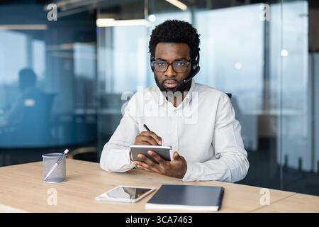 A businessperson with a headset working at a table, writing on a notepad, and using technology including a smartphone and tablet in a modern office environment. Stock Photo