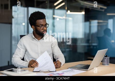Young professional man engaged in reviewing documents while seated at a desk in a modern office environment. He displays focus and dedication, utilizing a laptop and papers to complete his tasks. Stock Photo