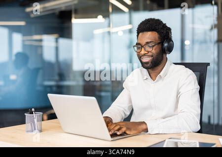A happy office worker wearing headphones, focused while working on a laptop in a contemporary workspace. The environment promotes productivity and comfort Stock Photo