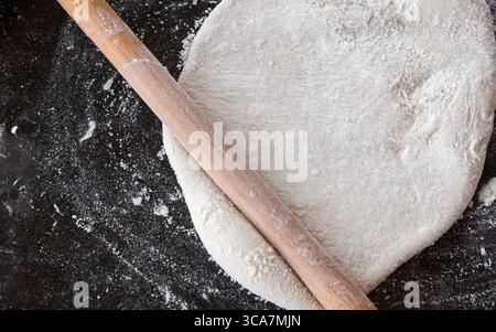 A close-up, top-down view of dough being flattened on a dark floured surface with a wooden rolling pin. Stock Photo