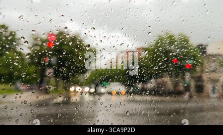 Raindrops on a car window create a soft, distorted view of city streets, traffic lights, and buildings on a rainy day. Stock Photo