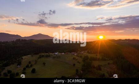 Golden sunset light casts a soft glow over rural hills and forests, with scattered houses nestled in open fields Stock Photo