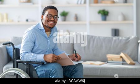 Happy with disability black guy in wheelchair taking notes, looking at camera and smiling at home, copy space Stock Photo